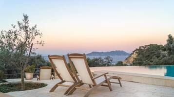 Un espace aménagé au bord de la piscine, avec deux chaises longues en bois et une petite table, offrant une vue dégagée sur une piscine aux eaux bleues et un paysage pittoresque de la mer et des montagnes lointaines, particulièrement beau au coucher du soleil.