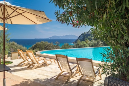 A poolside area with lounge chairs and an umbrella, overlooking a clear blue sea and distant mountains.