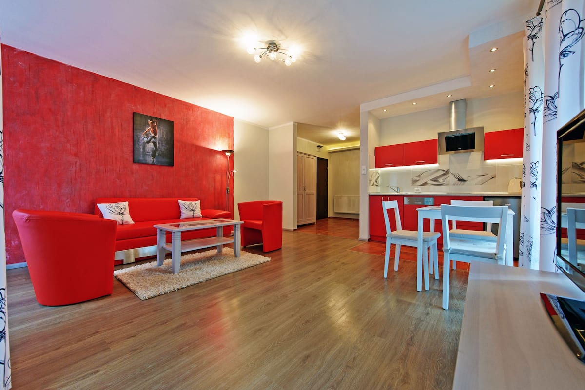 Ein modernes Wohnzimmer mit einer roten Couch und passenden Sesseln, ein Essbereich mit weißen Stühlen und eine Küche mit roten Schränken und einem eingebauten Backofen.