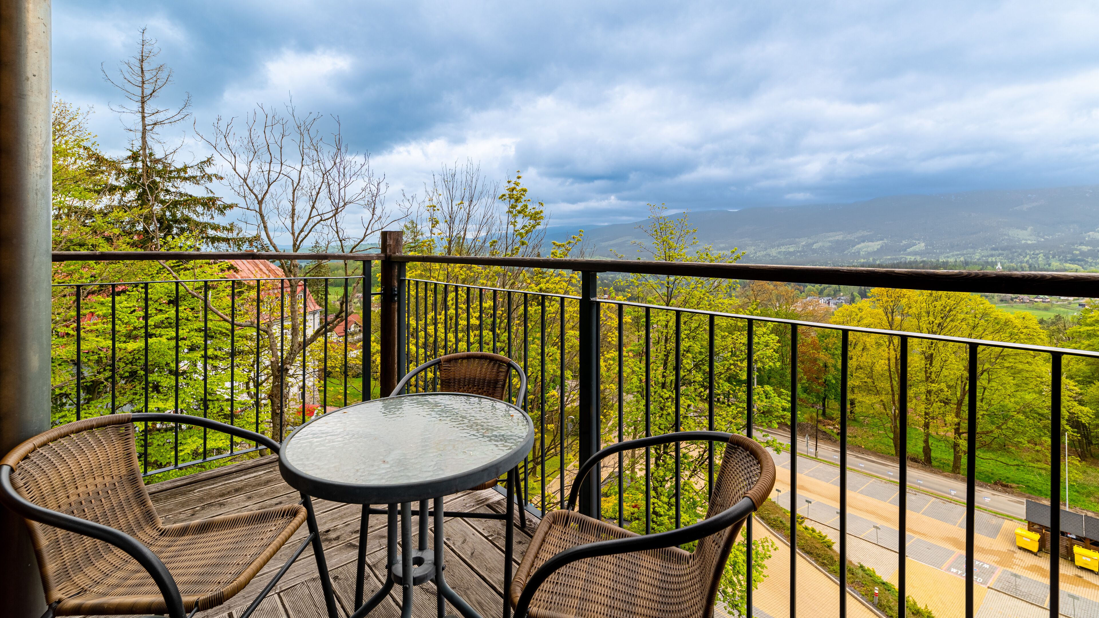 Een balkon met rieten stoelen en een glazen tafel, met uitzicht op een weg en groen.