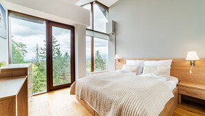 A bedroom with a large bed, wooden nightstands, a desk, and a view of trees through large windows. - Sun & Snow Resort Sudety (Szklarska Poreba)