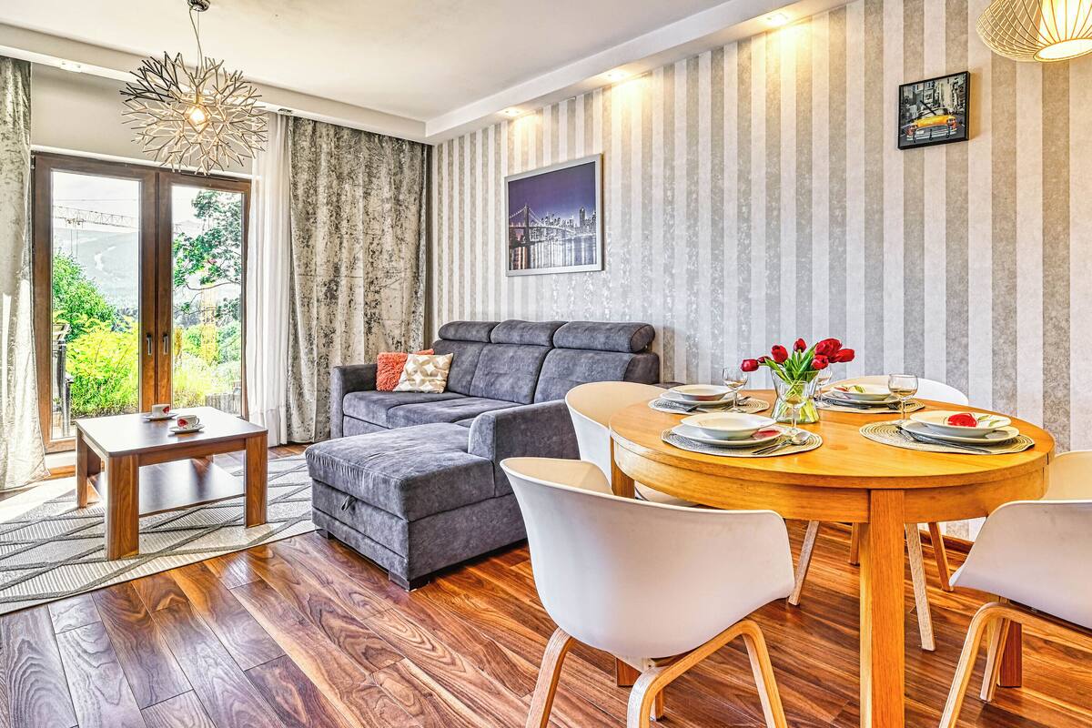 Ein modernes Wohnzimmer mit einem grauen Sofa, einem Holzes Esstisch für zwei Personen und einem Stadtbild an der Wand.