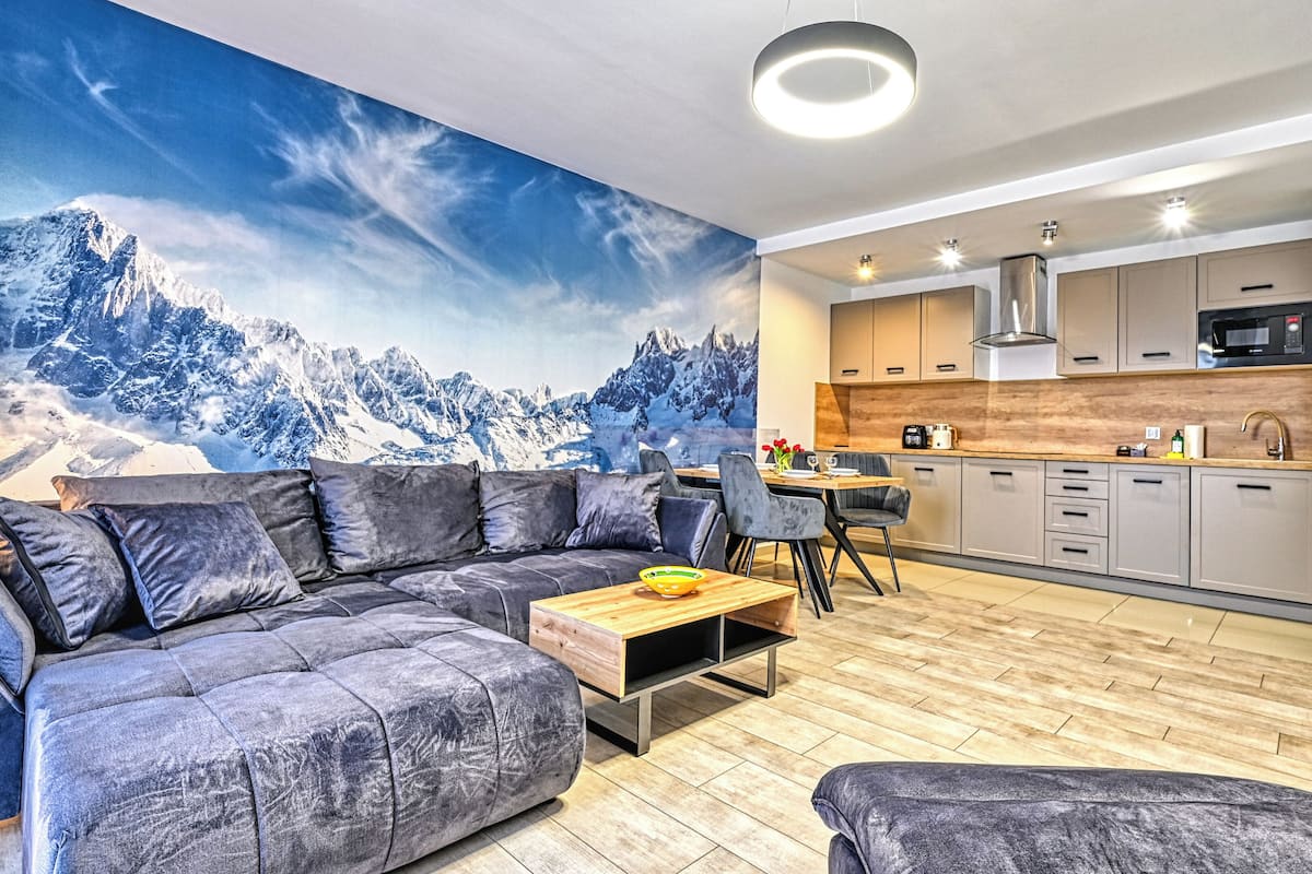 Ein modernes Wohnzimmer mit grauem Ecksofa, einem Holztisch und einer Wandmalerei mit Bergblick.