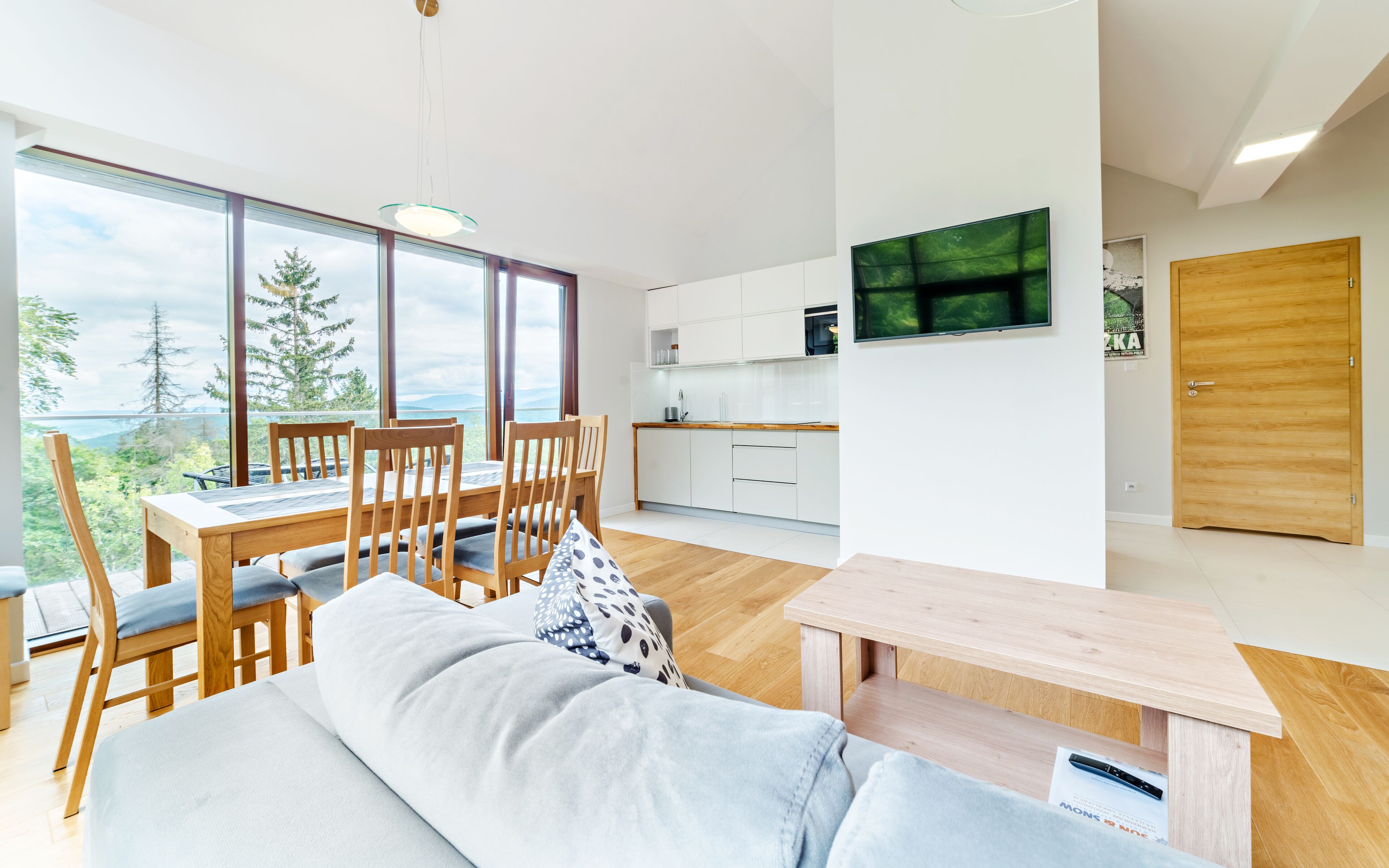Un salon moderne comprenant un canapé, une table à manger et des chaises, une cuisine avec des meubles blancs et une grande fenêtre donnant sur un paysage de arbres et de montagnes.