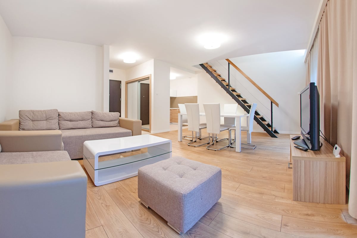 Ein modernes Wohnzimmer mit einer Couch, einem Hocker und einem Glastisch. Eine Treppe führt zu einem Essbereich mit weißen Stühlen und einem Tisch.