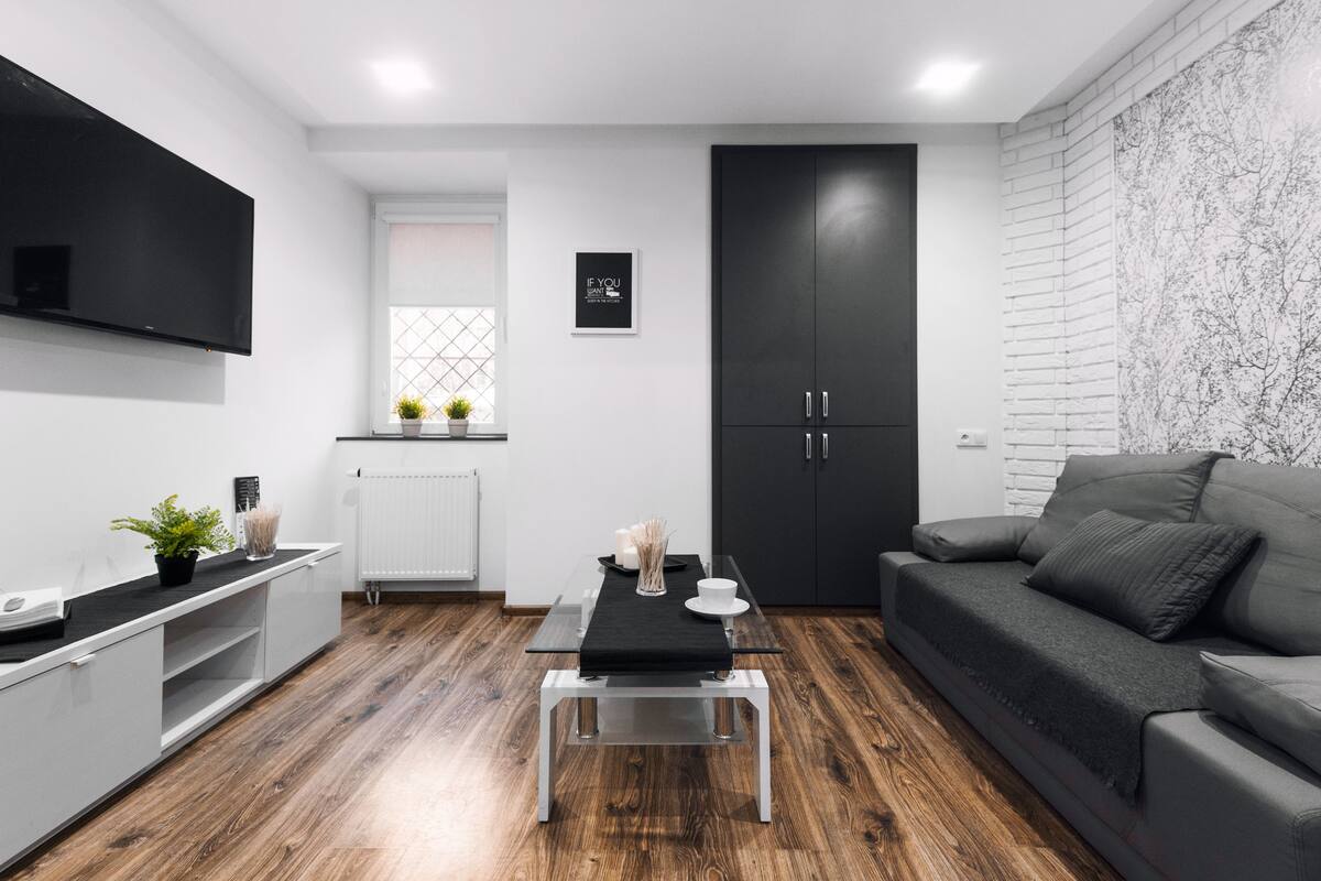 Ein modernes Wohnzimmer mit grauem Sofa, einem an der Wand befestigten schwarzen Fernseher, einem schwarzen Schrank und Holzboden.