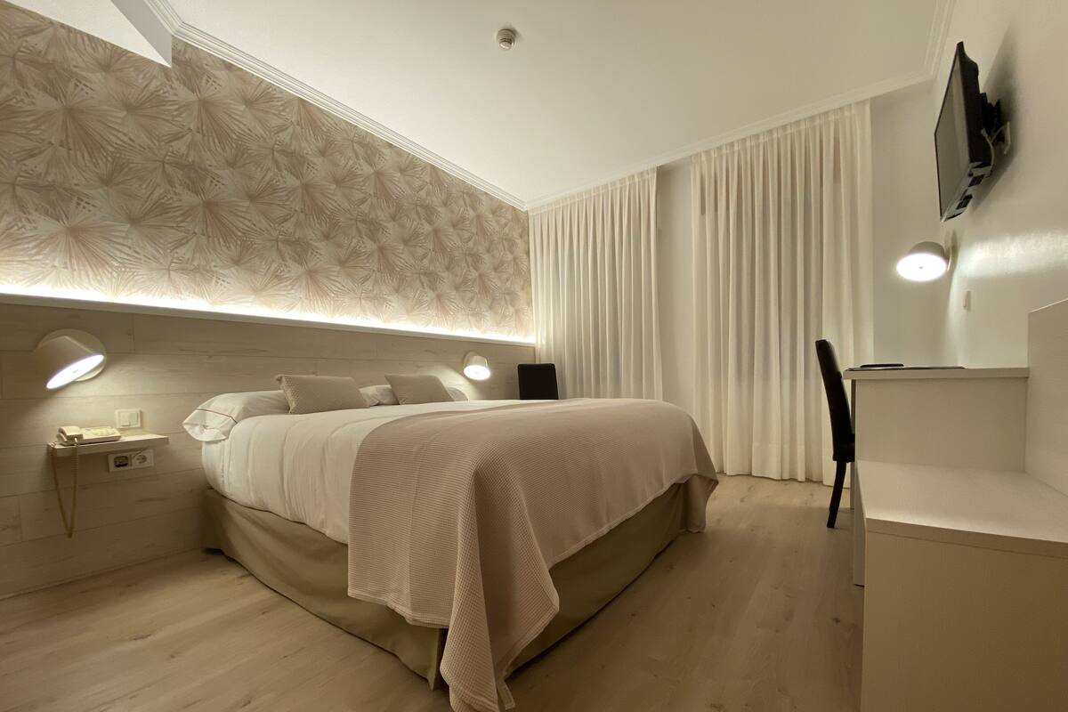 A modern bedroom with a large bed, bedside lamps, a desk, and a flat-screen TV mounted on the wall.