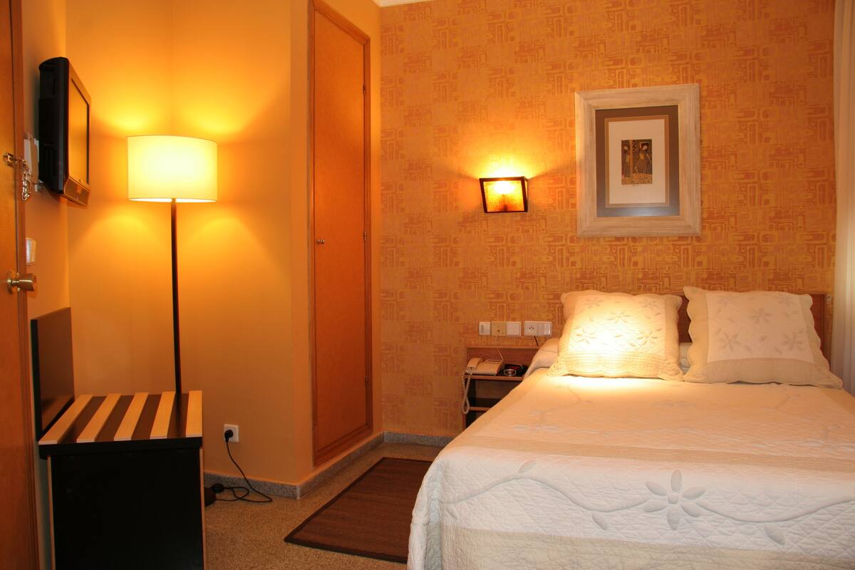 A hotel room with a bed, a nightstand, a lamp, a wall-mounted picture, and a door.