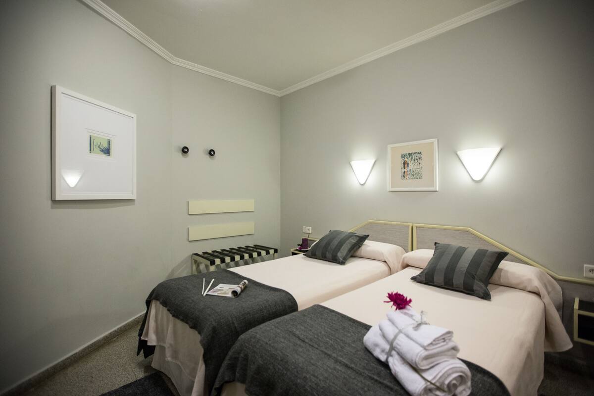 A hotel room with two beds, a small table, wall-mounted light fixtures, and framed artwork.