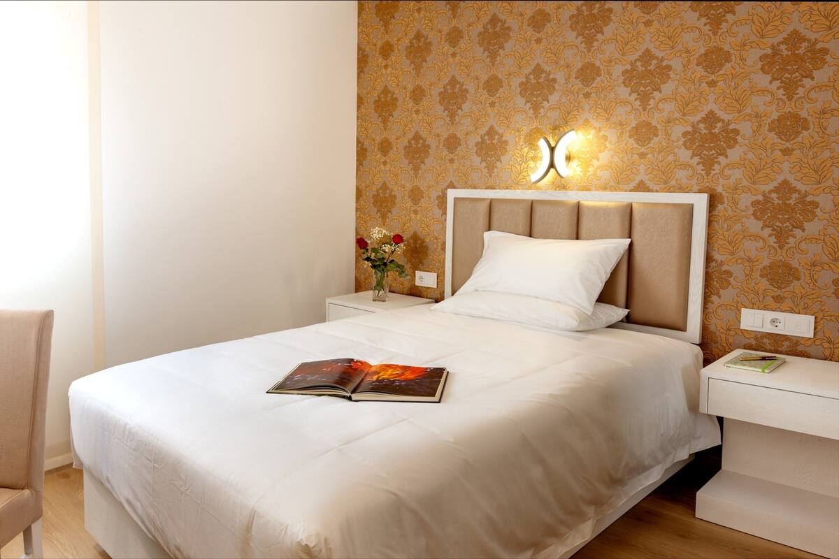 A neatly made bed with a white comforter and pillows, a bedside table with a book and a vase of flowers, and a wall with a floral pattern.