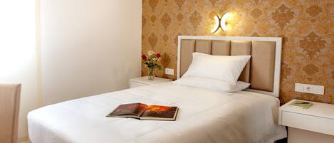 A neatly made bed with a white comforter and pillows, a bedside table with a book and a vase of flowers, and a wall with a floral pattern.