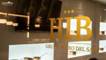 Un interior de hotel con un gran letrero 'HIB', un televisor montado en la pared y una fila de vasos en un estante.