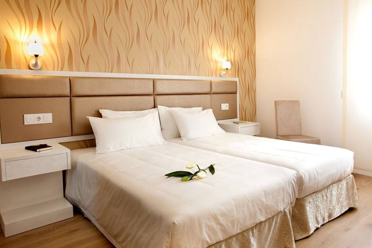 A hotel room with a large bed, two bedside tables, a nightstand, a wall-mounted lamp, and a decorative plant.