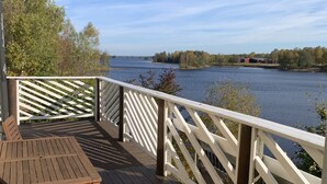 Een houten terras met een tafel en stoelen, met uitzicht op een rustig meer met bomen en gebouwen in de verte.