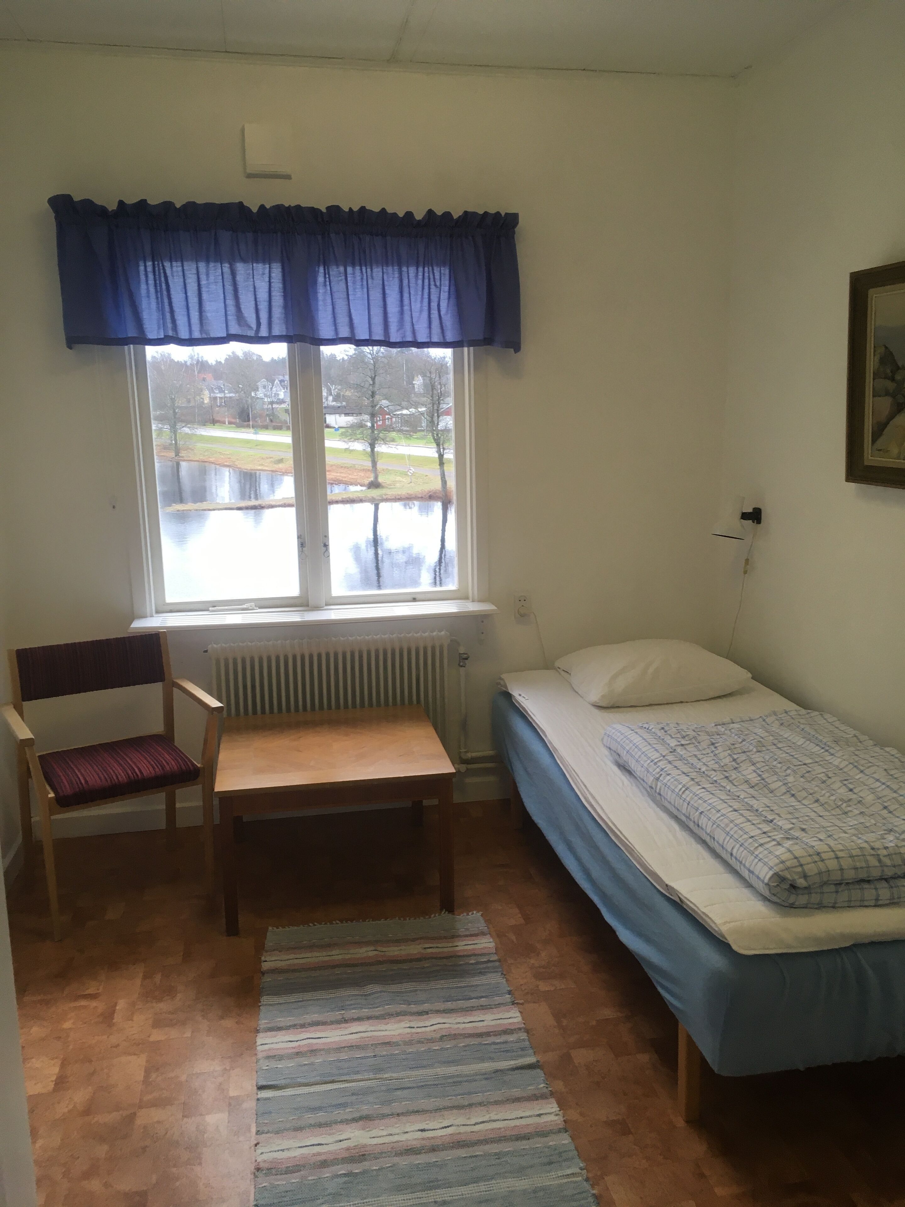 A small room with a single bed, a chair, a table, a radiator, and a window with a view of a body of water and houses.