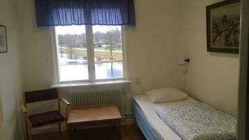 Una habitación pequeña con una cama individual, una silla, un radiador y una ventana que da a un parque y una masa de agua.