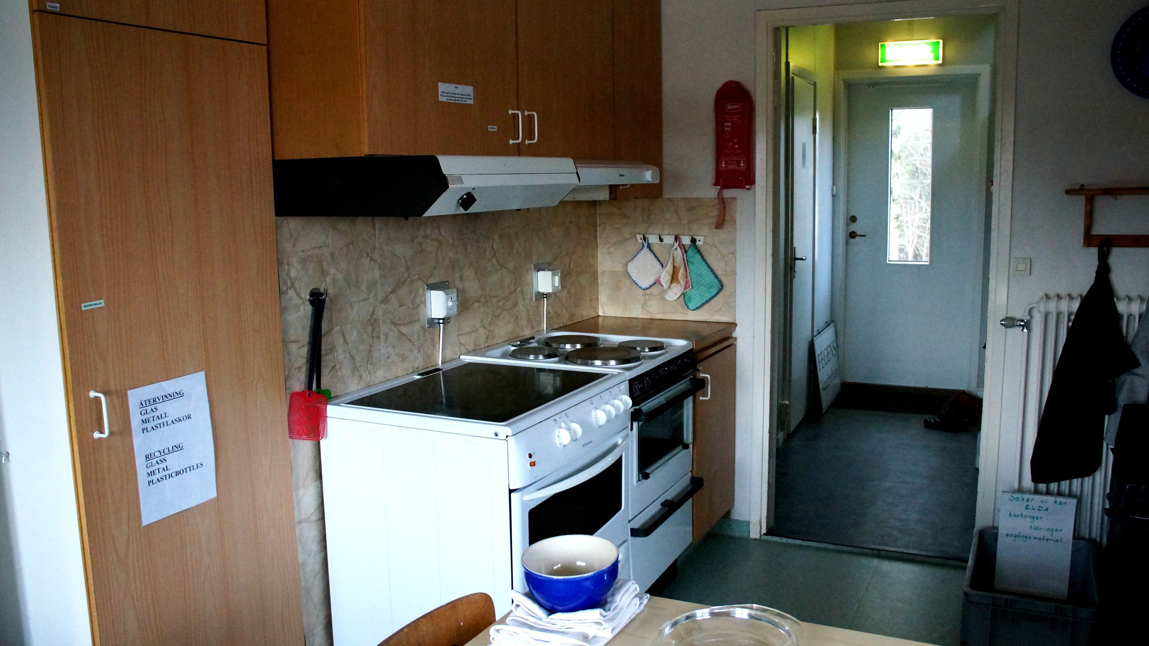 Cocina compacta con armarios de madera, estufa y horno. Señales indican zonas de reciclaje y gestión de residuos.
