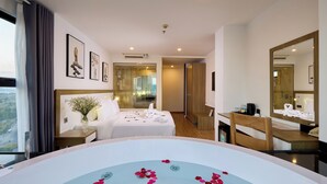 Habitación de hotel con una bañera grande llena de pétalos de rosa, una cama con sábanas blancas, un escritorio de madera y un espejo grande.