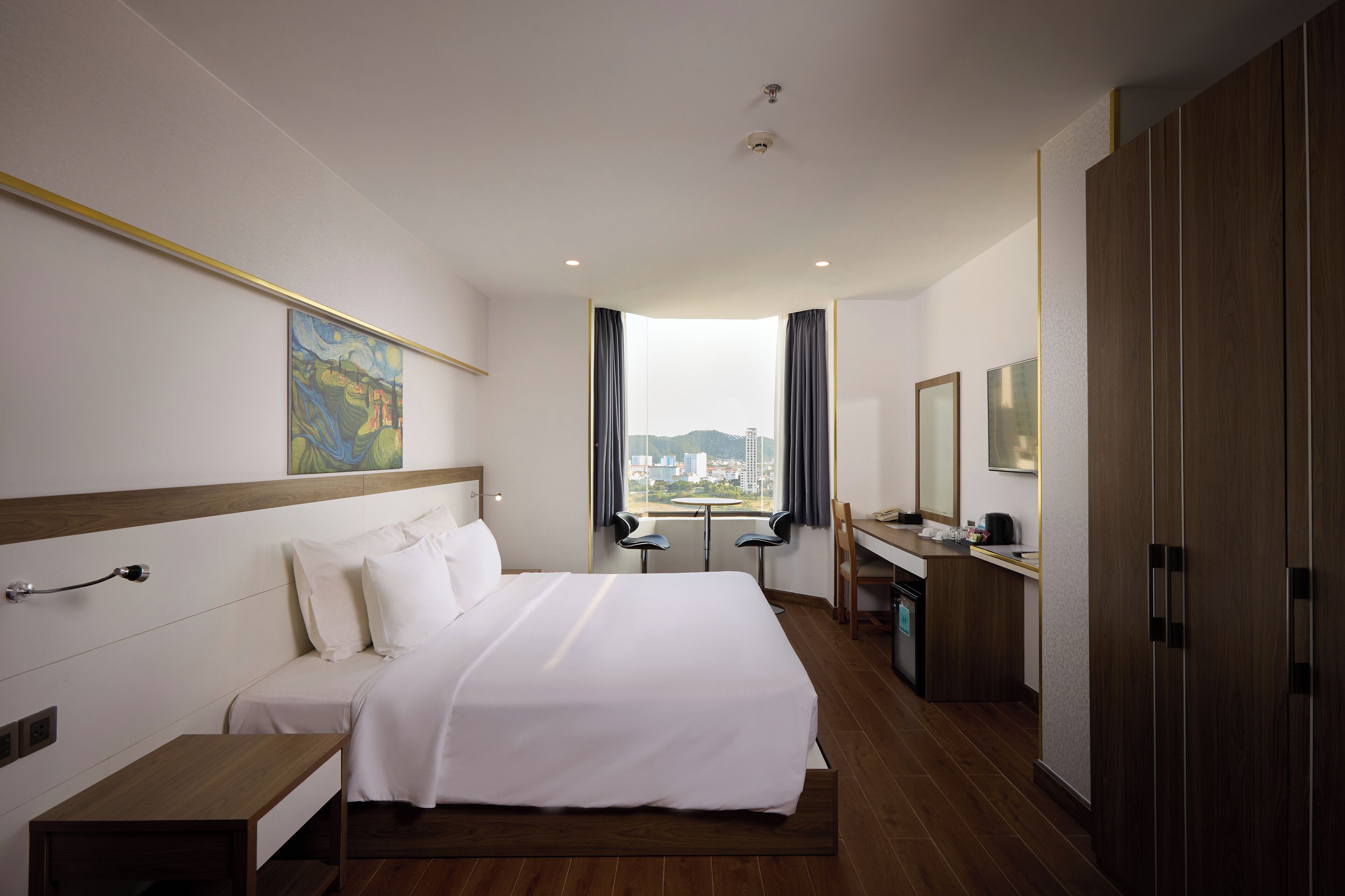 A hotel room with a large bed, a desk, a chair, and a view of the city.