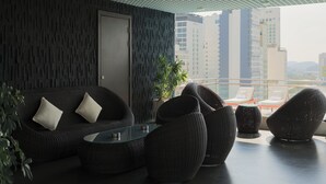 Un espacio lounge moderno con mobiliario de mimbre, una mesa de centro de vidrio y vistas a edificios altos.