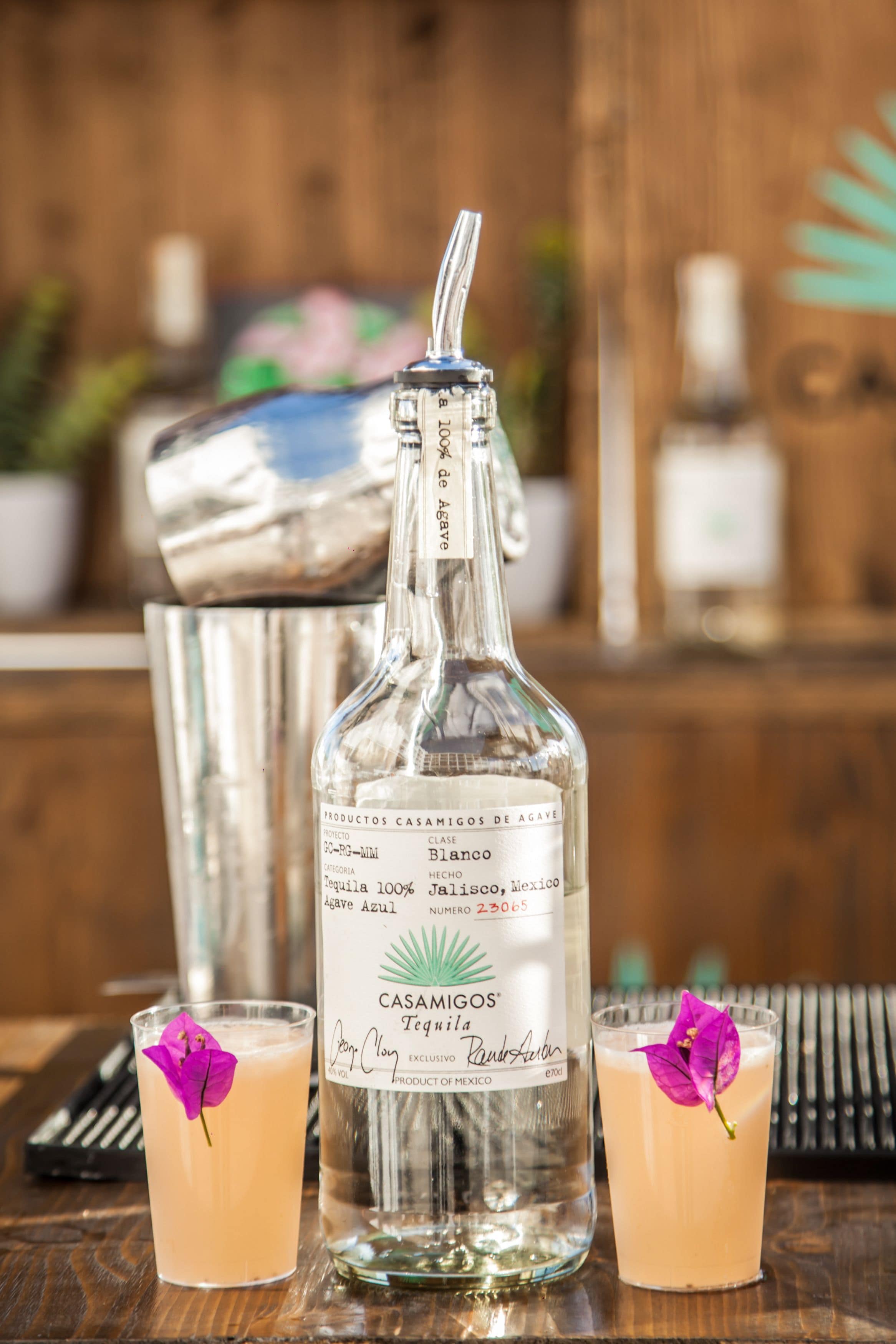A bottle of Casamigos Tequila with a silver cap and a straw, two glasses of a light-colored cocktail with purple flowers, and a metal shaker in the background.