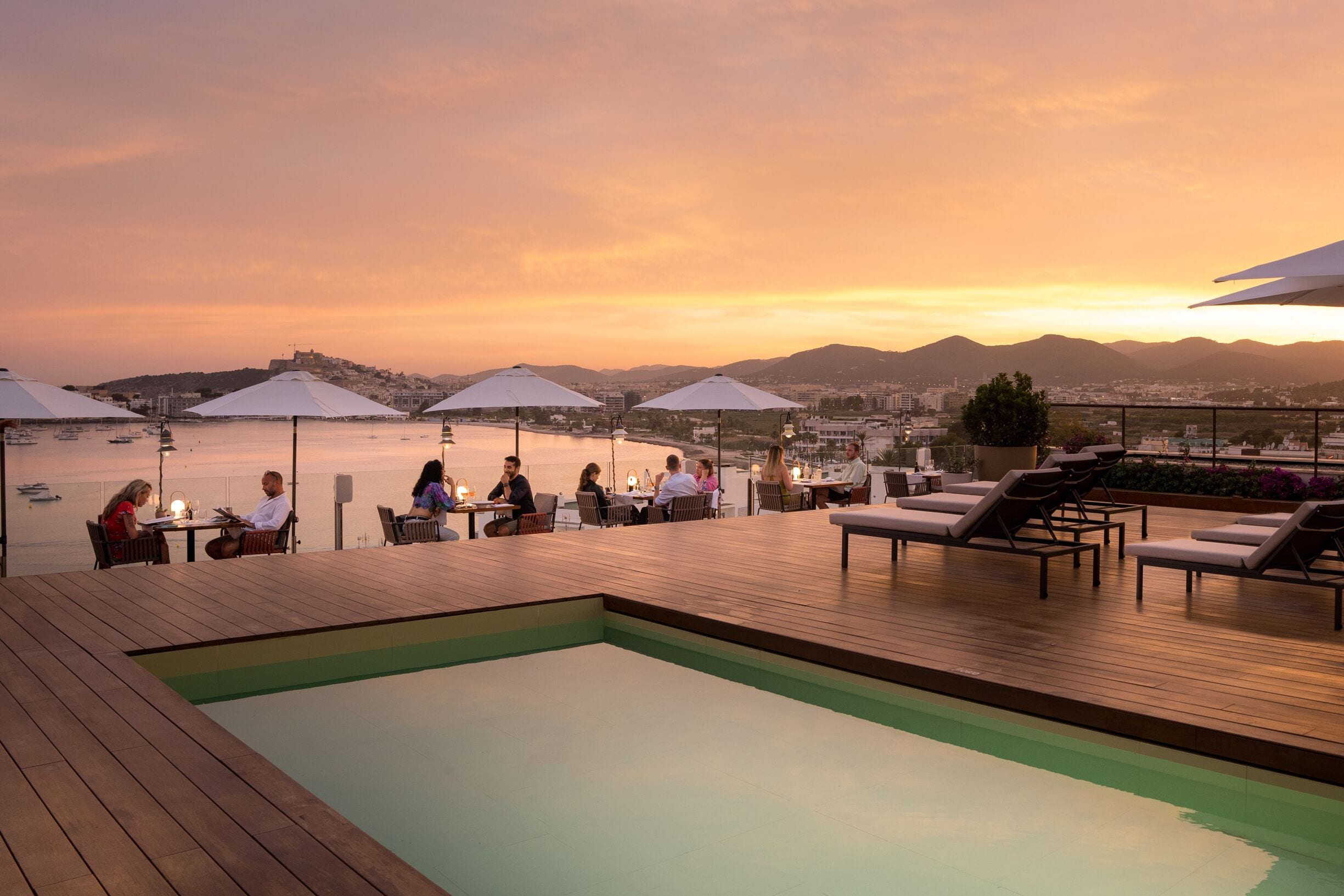 A rooftop pool area with lounge chairs and umbrellas, overlooking a body of water and cityscape during sunset.