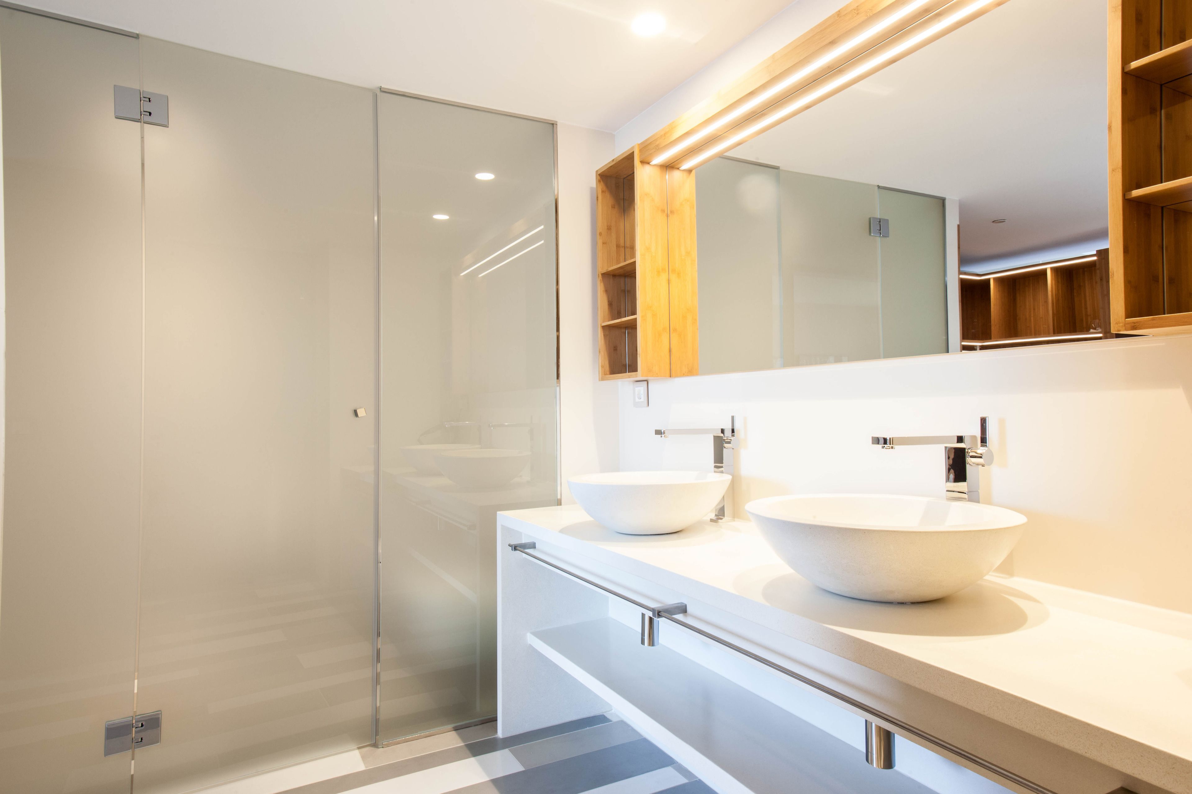 Un baño moderno con cabina de ducha de vidrio, dos lavamanos sobre encimera y un mueble de tocador de madera.