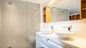 Un baño moderno con cabina de ducha de vidrio, dos lavamanos sobre encimera y un mueble de tocador de madera.