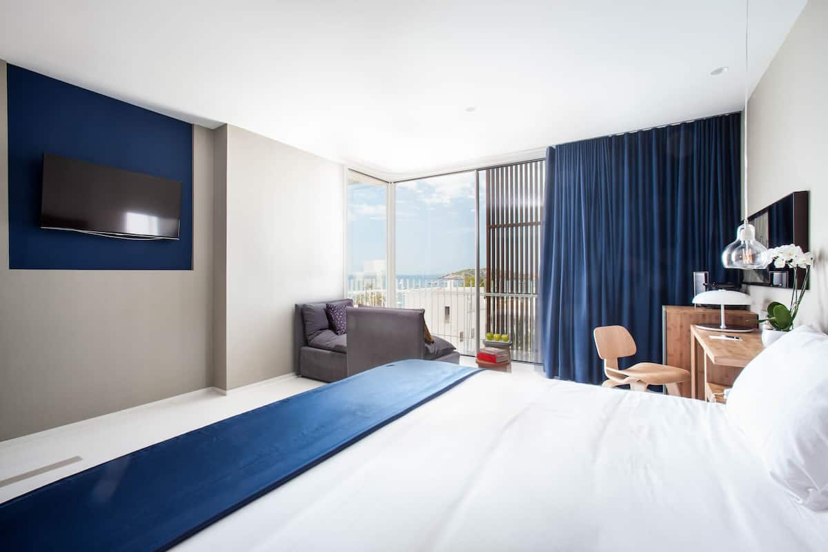 A modern hotel room with a large bed, a desk, a chair, a TV, and a view of the city.