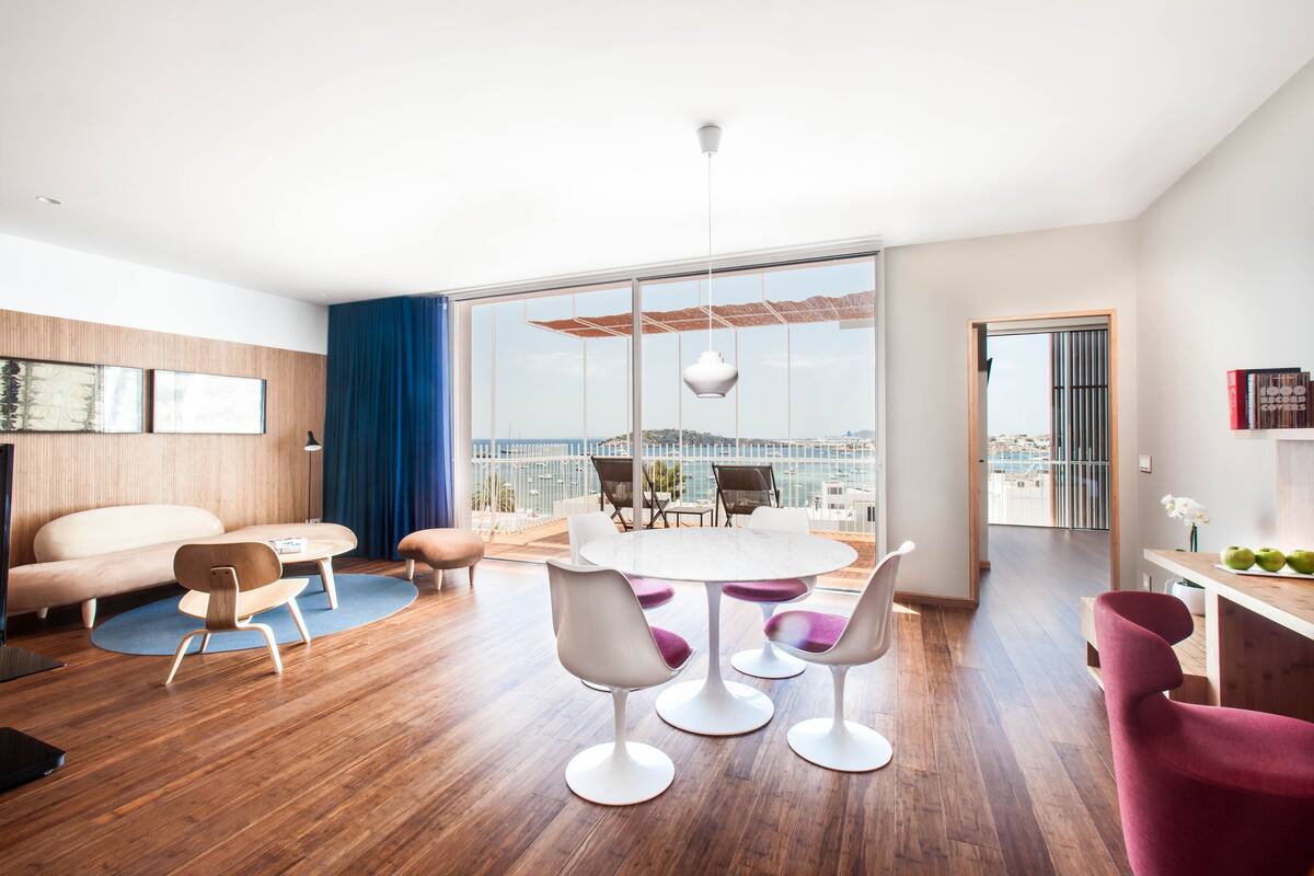 A modern living room with a wooden floor, a round dining table, and a large window offering a view of the city.