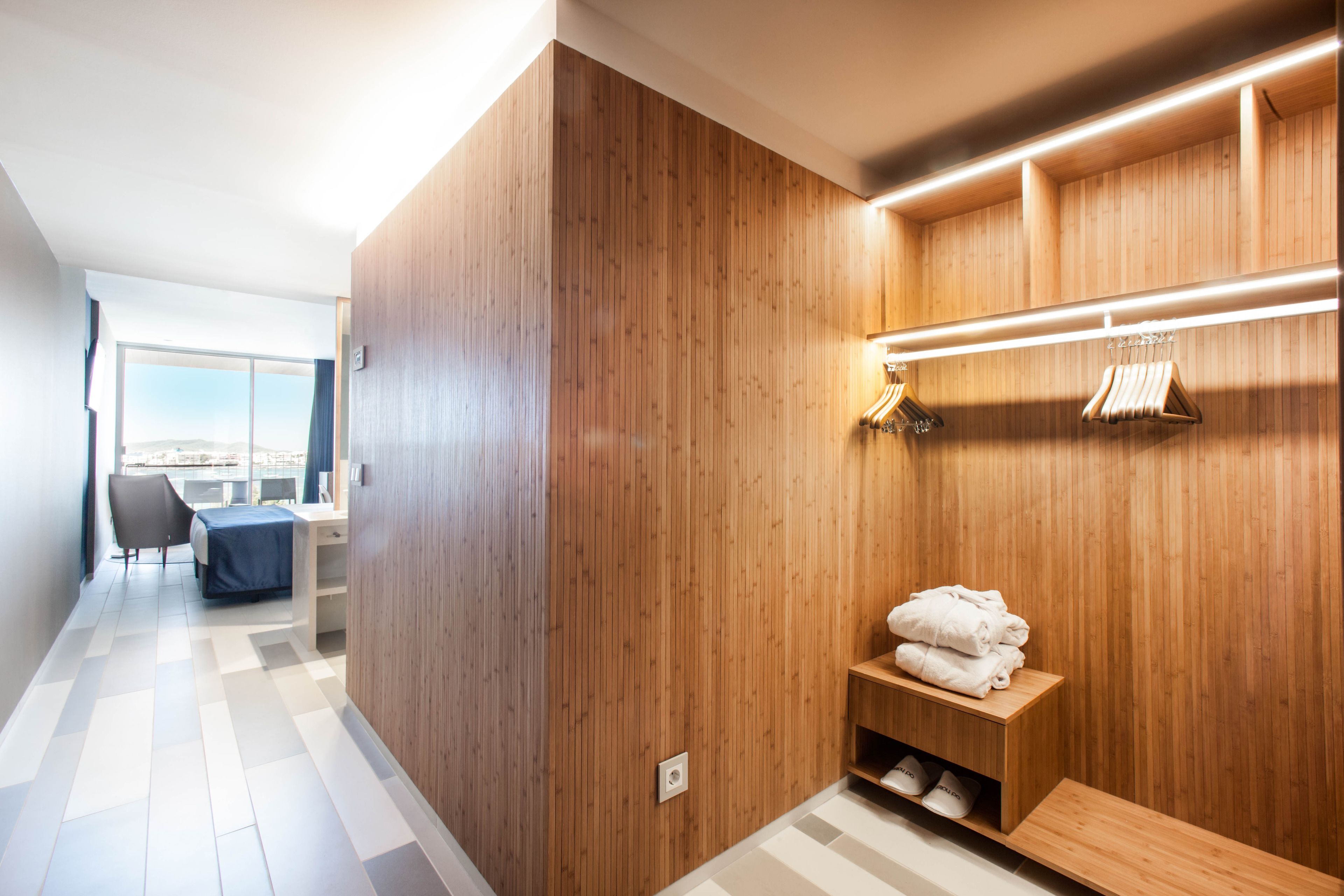 A modern hotel room with wooden panel walls, a built-in bench with towels, and a view through a large window.