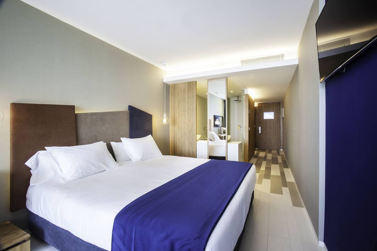 A hotel room with a large bed, a bedside table, a mirror, and a bathroom with a sink and mirror.