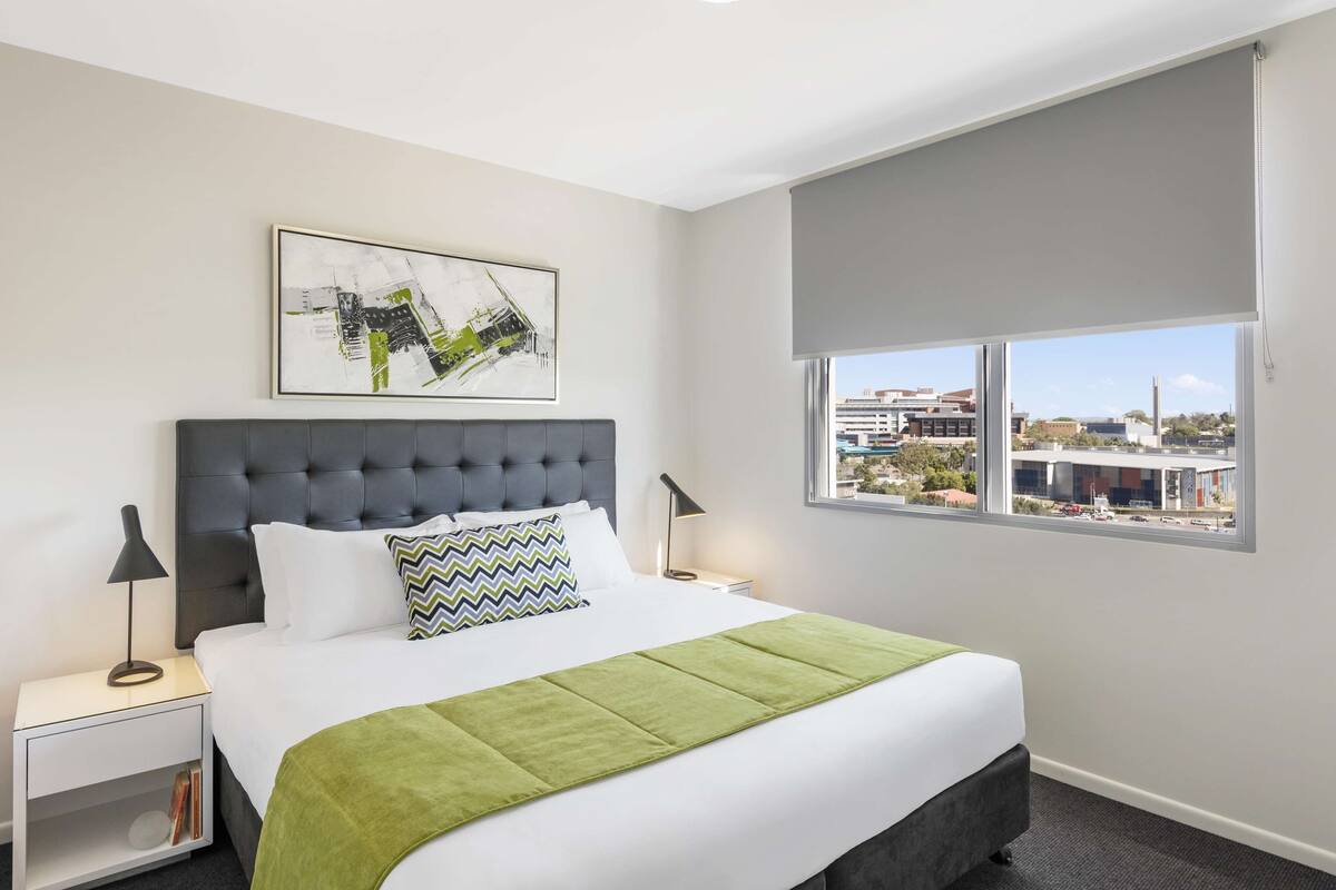 Une chambre d’hôtel avec un lit, une table de chevet, une lampe, un plaid vert, une vue sur la ville et un tableau encadré au mur.