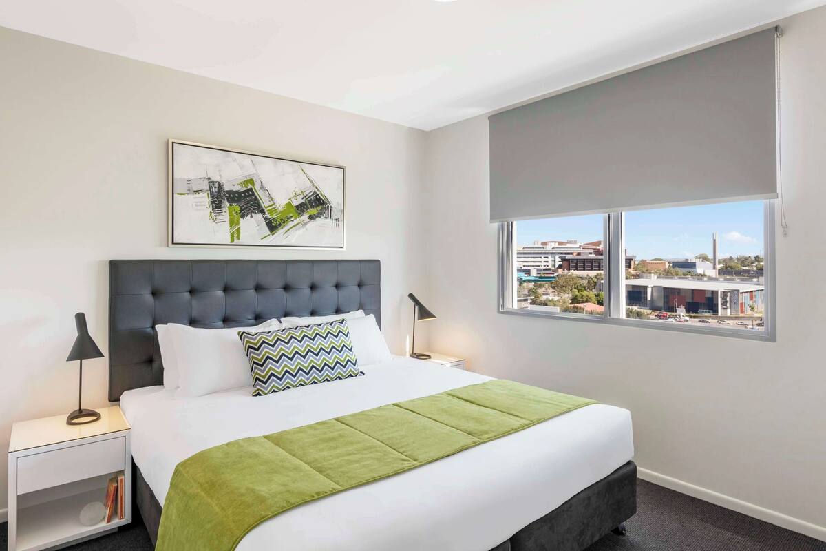 Une chambre d’hôtel avec un grand lit, une vue sur la ville, une tête de lit grise et un tableau encadré au mur.