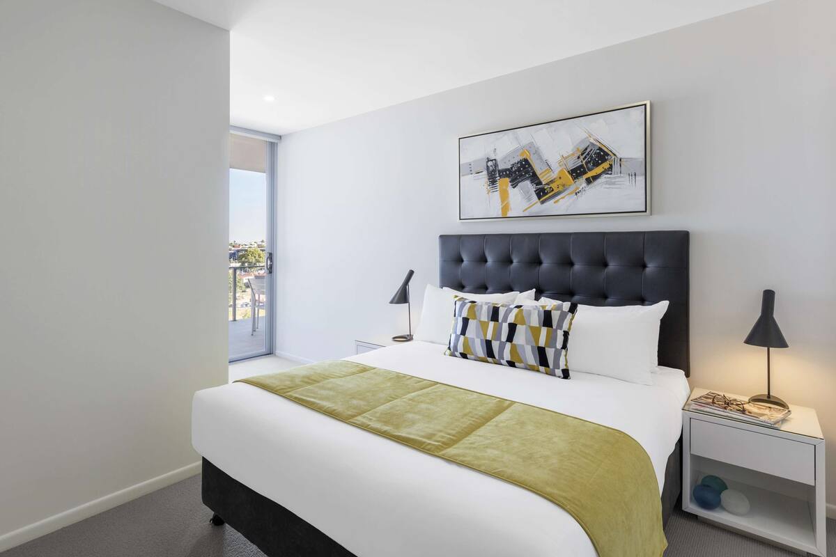 Une chambre d’hôtel moderne avec un grand lit, une vue sur l’extérieur, une œuvre d’art encadrée au mur et une table de chevet avec une lampe.