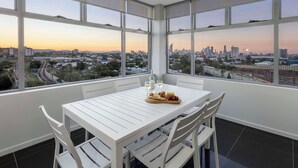 Un coin repas avec une table dressée pour deux personnes, de grandes fenêtres donnant sur la ville et une bouteille d’eau posée sur la table.