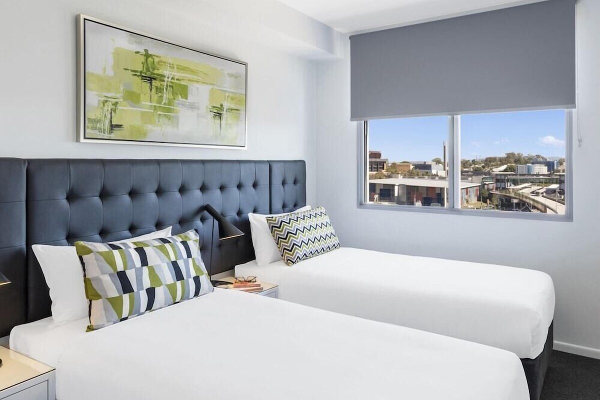 Habitación de hotel con dos camas, un ventanal con vistas, cabecera gris y un cuadro abstracto enmarcado.
