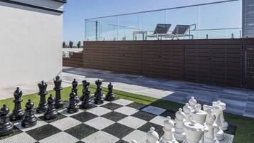 A checkered outdoor chessboard with chess pieces, a glass fence, and a building in the background.