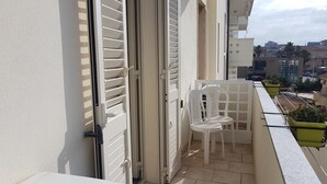 En balkon med en hvid stol og et lille bord, en hvid dør med skodder, og udsigt over bybilledet.