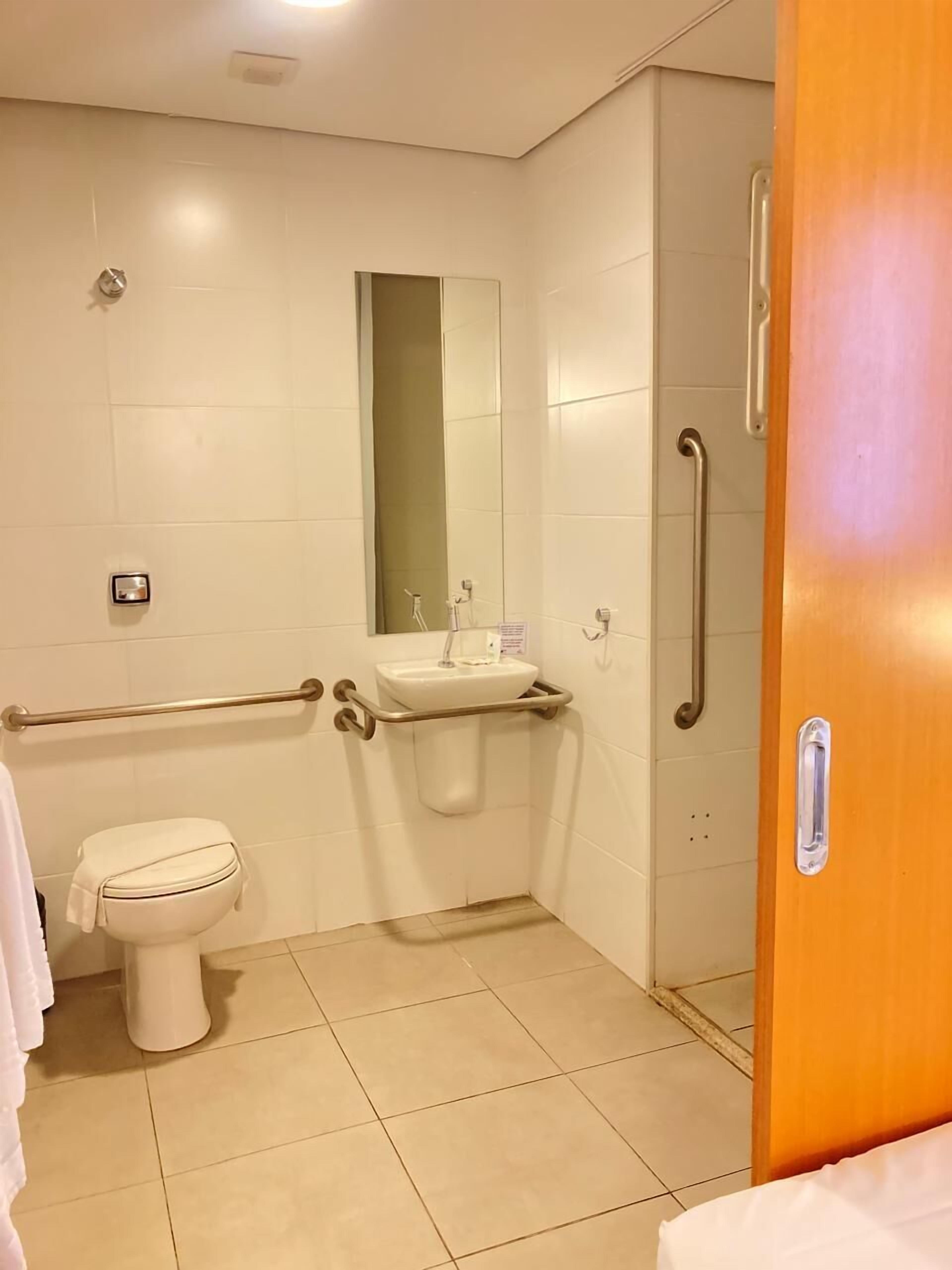 Baño con inodoro, lavamanos, espejo y barras de apoyo.