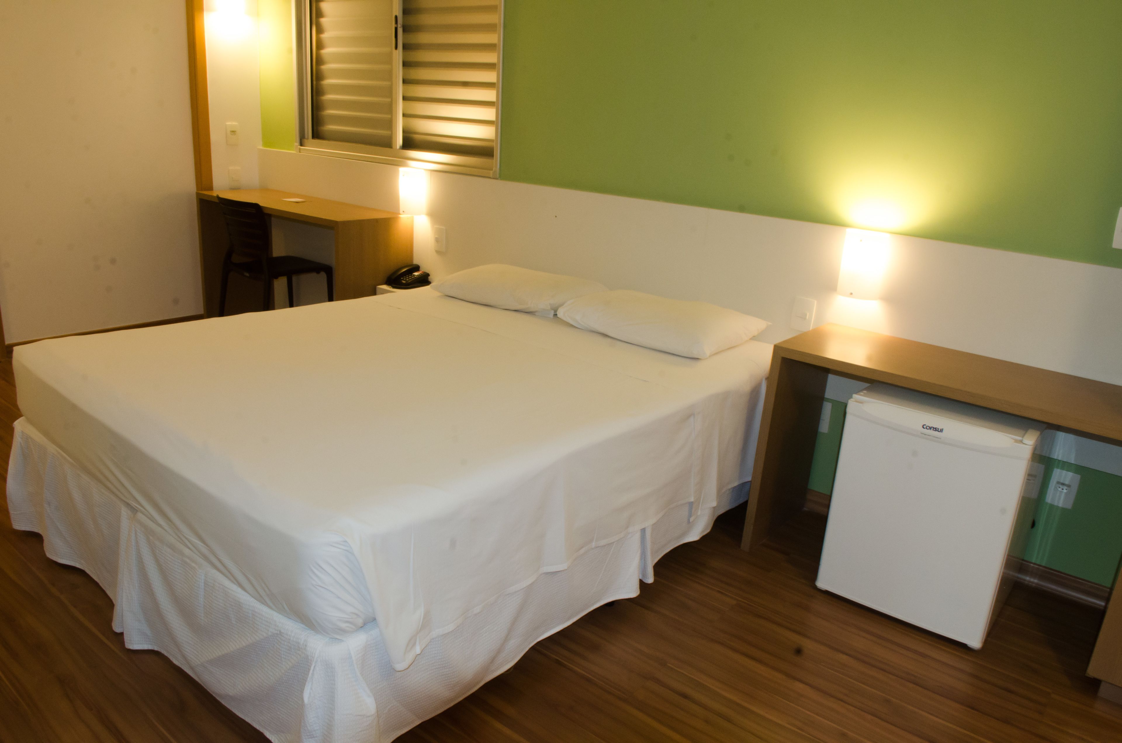 Habitación de hotel con una cama grande, un escritorio con silla, un refrigerador pequeño y una mesita de noche con lámpara.