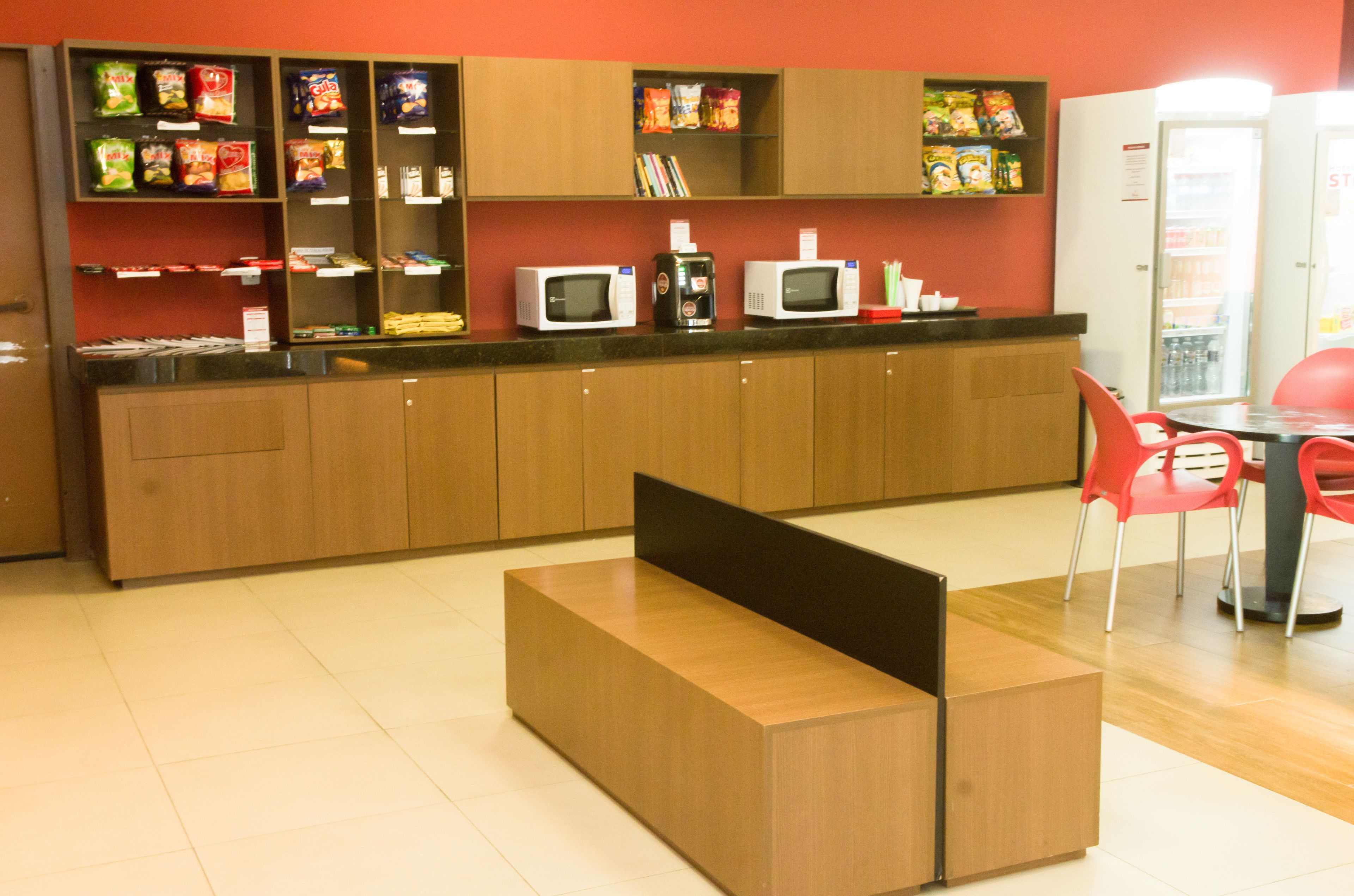 A modern kitchen area with wooden cabinets, a countertop with microwaves, and a seating area with red chairs.