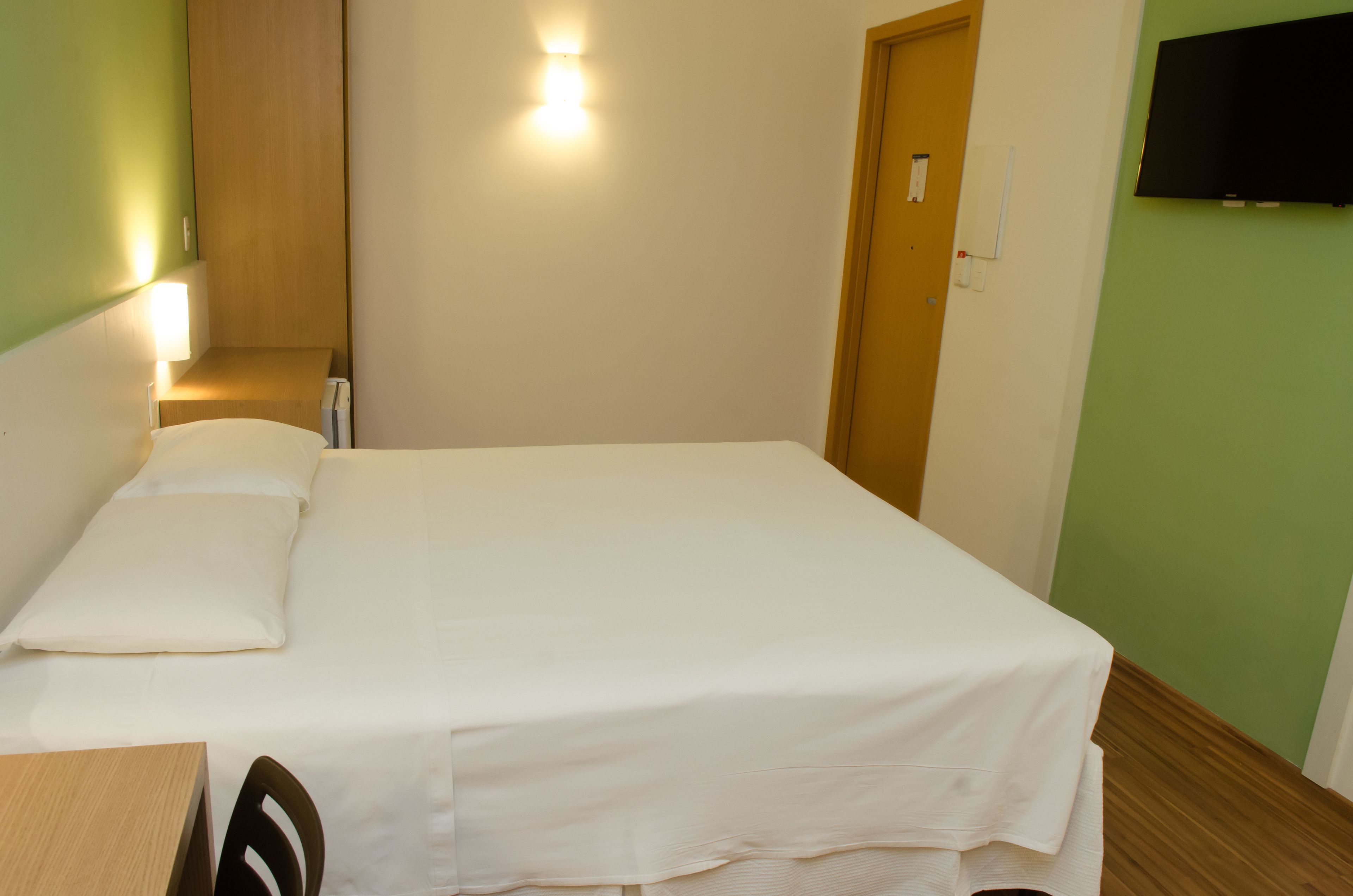 A single-bed room with a wooden headboard, a bedside table, a wall-mounted TV, and a green accent wall.
