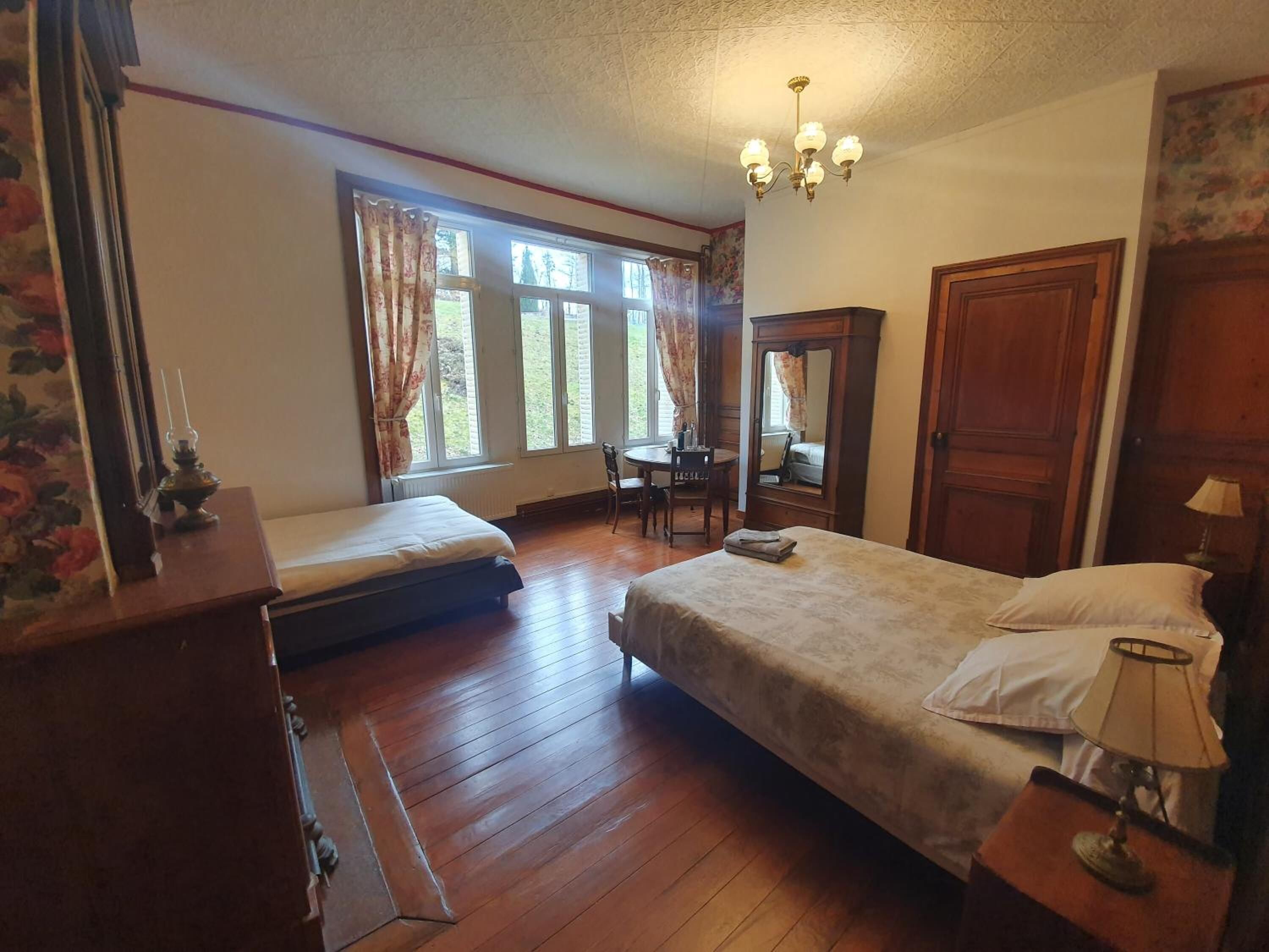 A bedroom with two beds, a wooden floor, a chandelier, a desk, and a window with curtains.