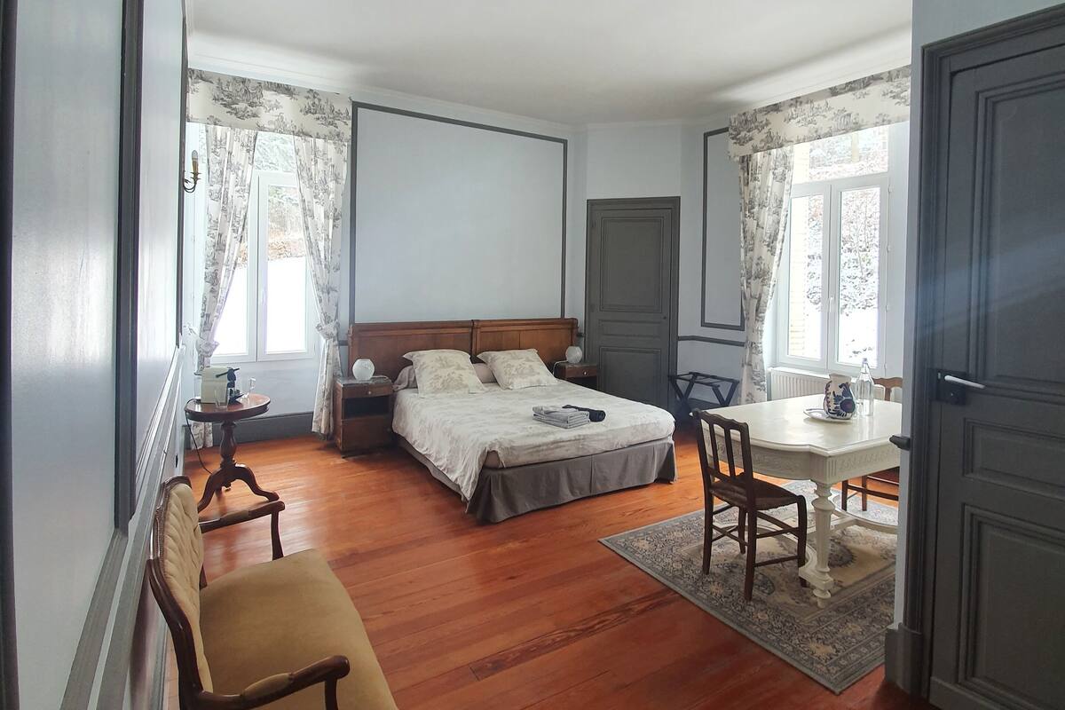 Un dormitorio con piso de madera, una cama con cubrecama blanco, una mesita de noche con lámpara, una silla y una pequeña mesa con jarrón y taza.