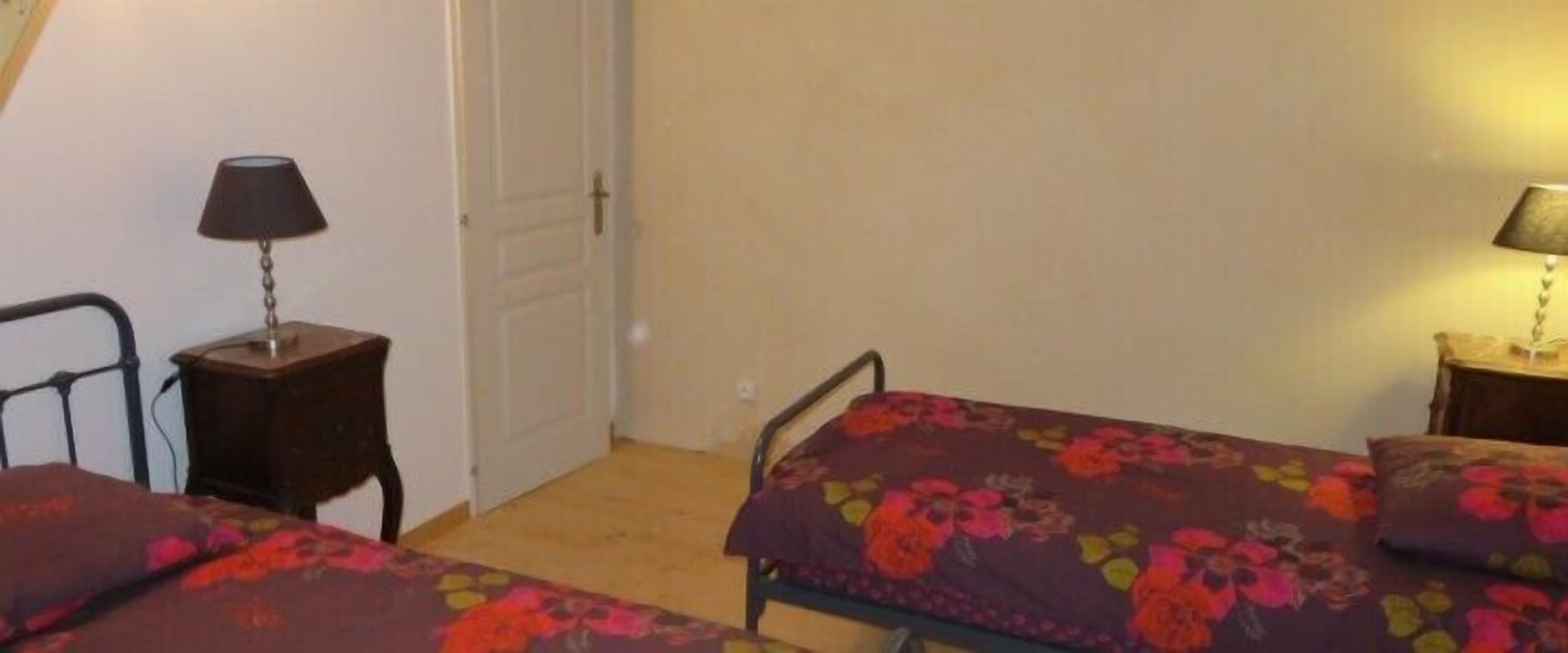Une petite pièce simple, avec deux lits simples, chacun recouvert d’une couvre-lit à motifs floraux. On y trouve deux tables de chevet avec des lampes, et une porte est visible au fond.