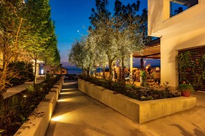 A nighttime outdoor area with a pathway, trees, and a seating area with tables and chairs. - Hotel Maximilian (Bardolino)
