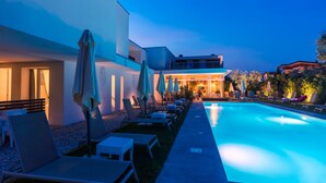 A swimming pool area with lounge chairs and umbrellas, a modern building with a glass facade, and a well-lit outdoor space. - Hotel Maximilian (Bardolino)