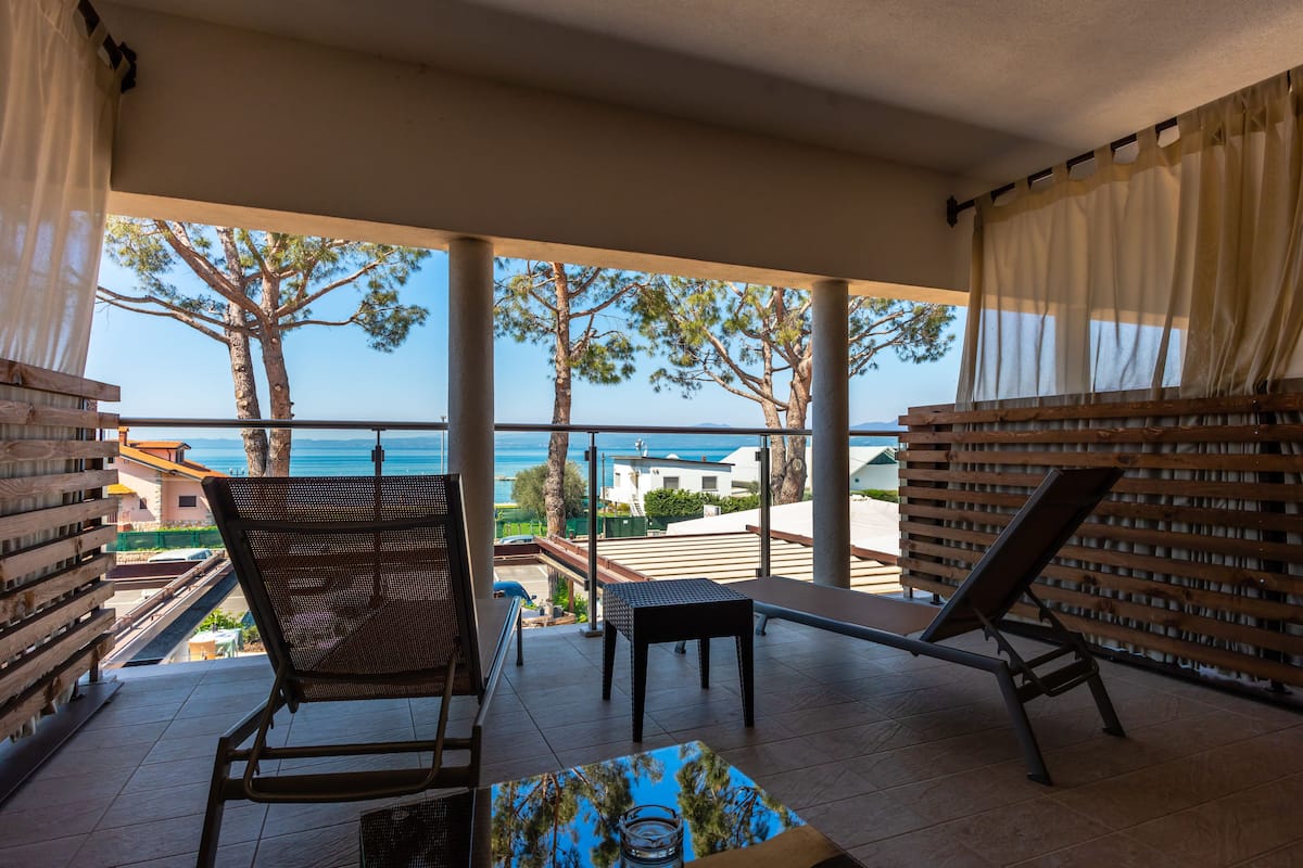 A balcony with two chairs, a small table, and a view of the sea and buildings.
