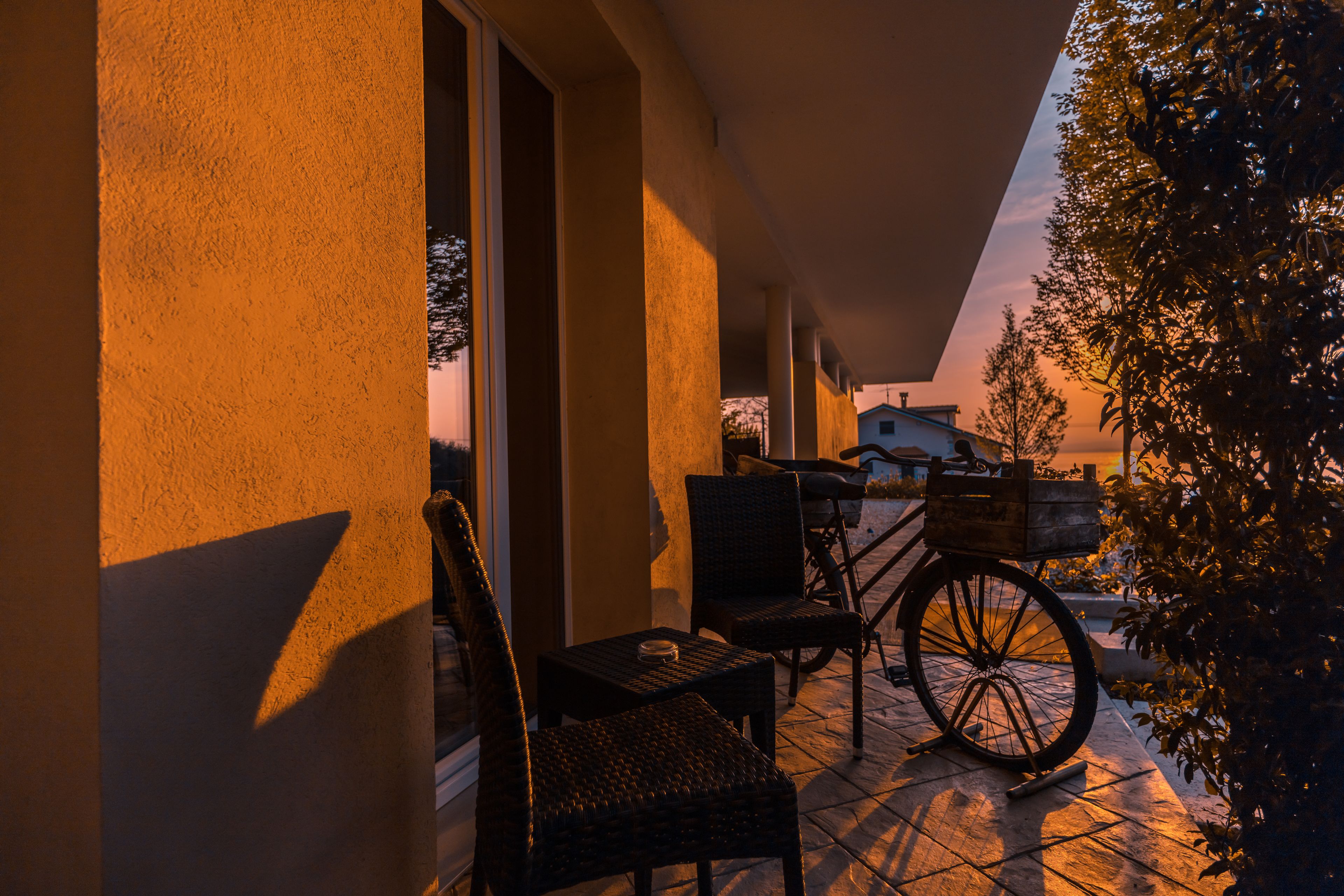 A patio with wicker furniture and a bicycle, a building with a sloped roof, and a tree in the background.